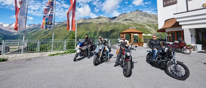 Four motorcyclists in front of Alpenhotel Laurin with mountains and flags
