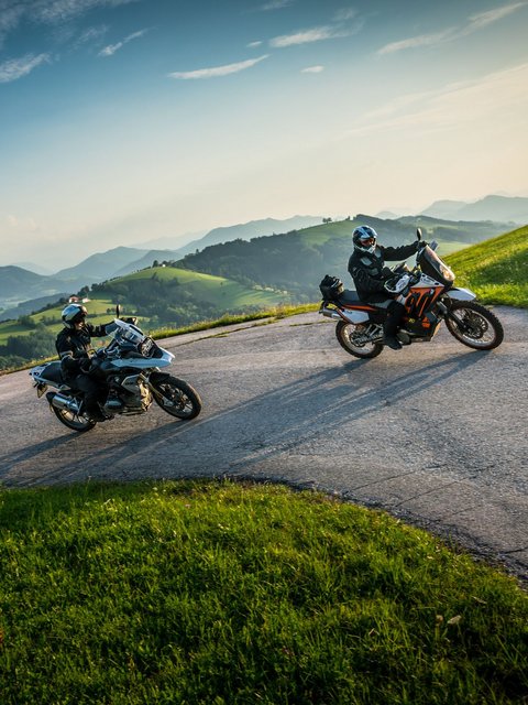 Zwei Motorradfahrer auf kurviger Bergstraße mit grünen Hügeln und blauem Himmel