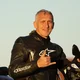 Man in leather motorcycle suit making hand gesture next to motorcycle