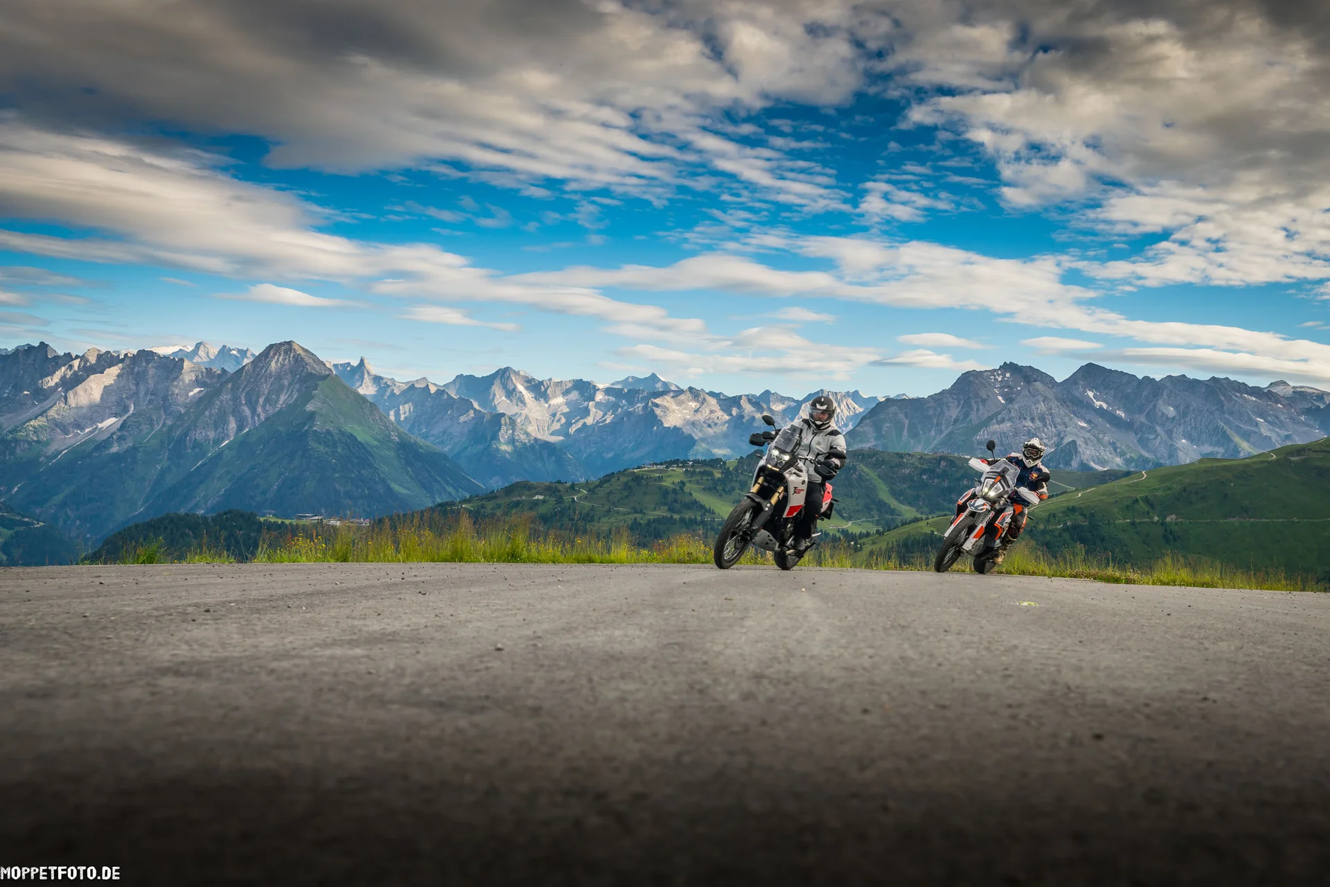 Zwei Motorradfahrer auf Bergstraße vor Alpenlandschaft unter bewölktem Himmel