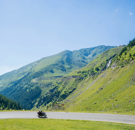 Motorradfahrer auf kurviger Bergstraße mit grünen Hügeln und blauem Himmel