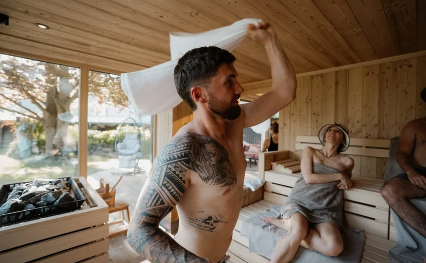 Sweaty Weekends Mann mit Tattoo wedelt Handtuch in Sauna mit anderen Gästen