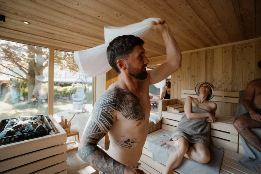 Mann mit Tattoo wedelt Handtuch in Sauna mit anderen Gästen