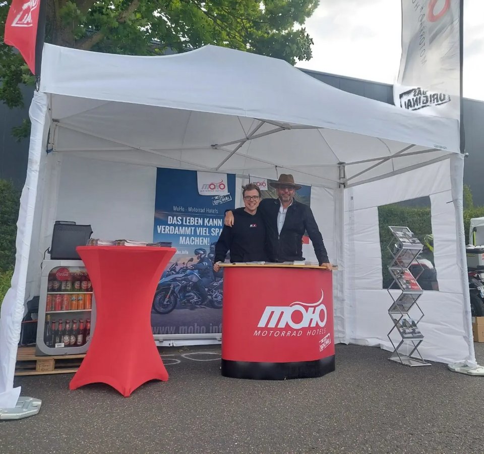 Zwei Personen an einem MoHo Motorrad Hotels Stand unter weißem Zelt