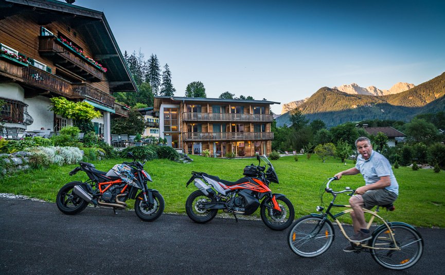 Mann auf Fahrrad vor zwei Motorrädern vor Alpenhäusern und Bergen