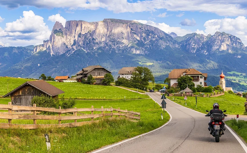 Motorradfahrer auf Landstraße mit Alpen und Bauernhöfen im Hintergrund