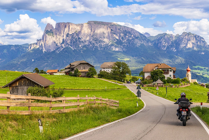 Motorradfahrer auf Landstraße mit Alpen und Bauernhöfen im Hintergrund