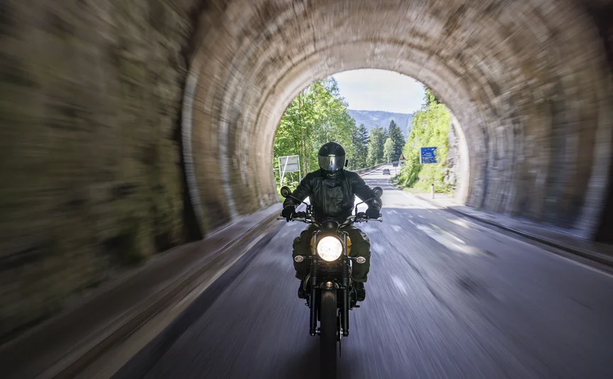 Gasthof Hotel Zur Post Samerberg *** © Detlef Louis Motorrad Vertriebs-GmbH Motorradfahrer fährt durch einen steinernen Tunnel, dahinter sind Bäume und Berge zu sehen
