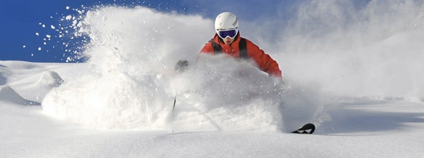 Skifahrer in roter Jacke fährt durch tiefen Pulverschnee bei klarem Himmel