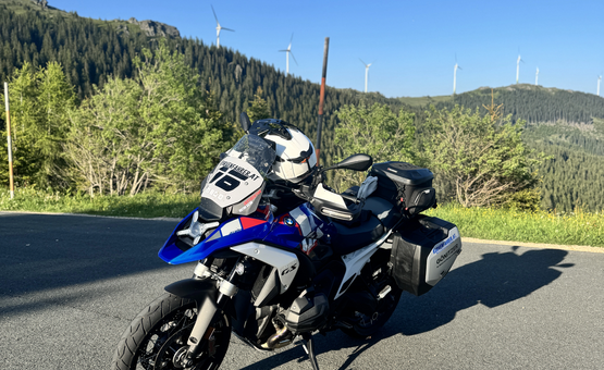 Motorrad steht auf Straße vor bewaldeten Hügeln und Windrädern bei klarem Himmel