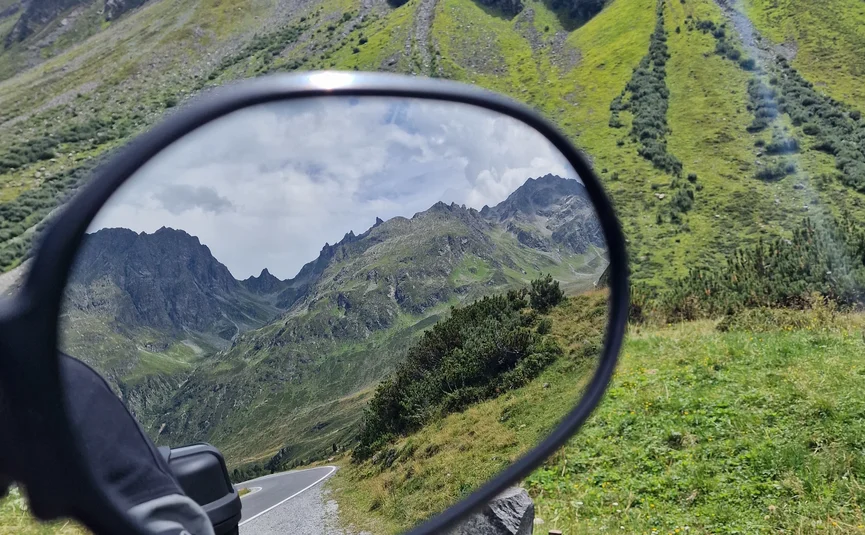View in motorcycle mirror reflecting mountain road and surrounding peaks