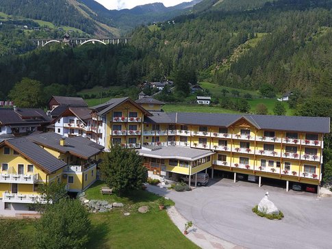 Erlebnishotel Kärnten-Mölltal © Erlebnishotel Kärnten Mölltal Gelbes Hotelgebäude in grüner Berglandschaft mit Autobahnbrücke im Hintergrund