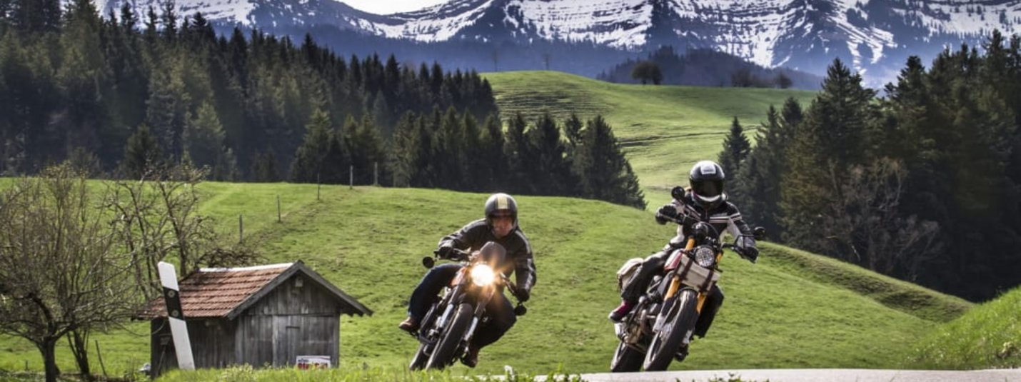 Zwei Motorradfahrer fahren auf einer Landstraße vor schneebedeckten Bergen.