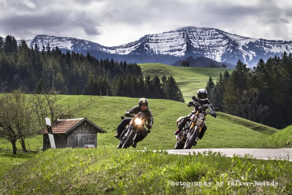 Two motorcyclists riding on a country road with snowy mountains in the background.
