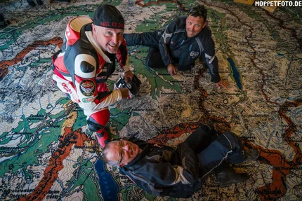 Three men in motorcycle gear on a large map spread on the floor