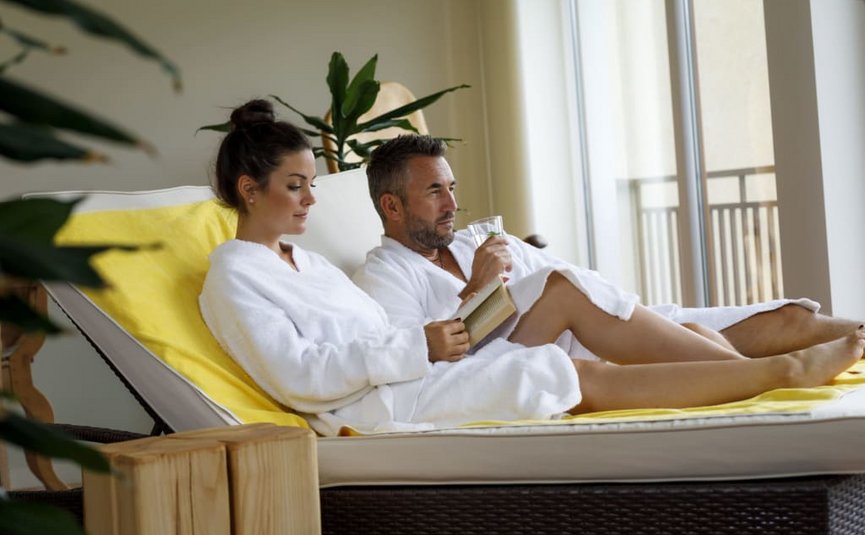 Paar entspannt auf Liege im Spa, Frau liest, Mann trinkt Wasser