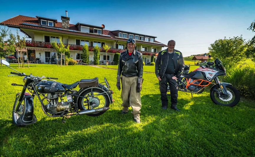 Two men with vintage and modern motorcycles on a lawn in front of a house