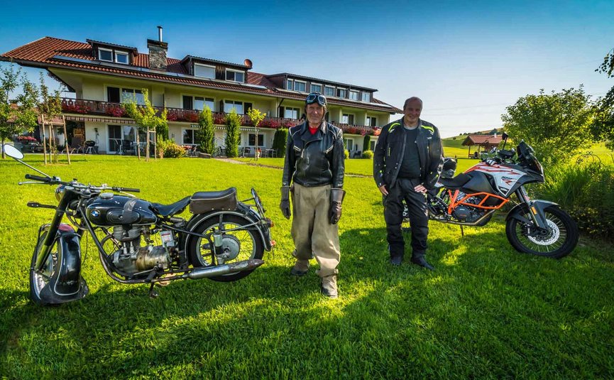 Zwei Männer mit Vintage- und modernen Motorrädern auf einer Wiese vor einem Haus