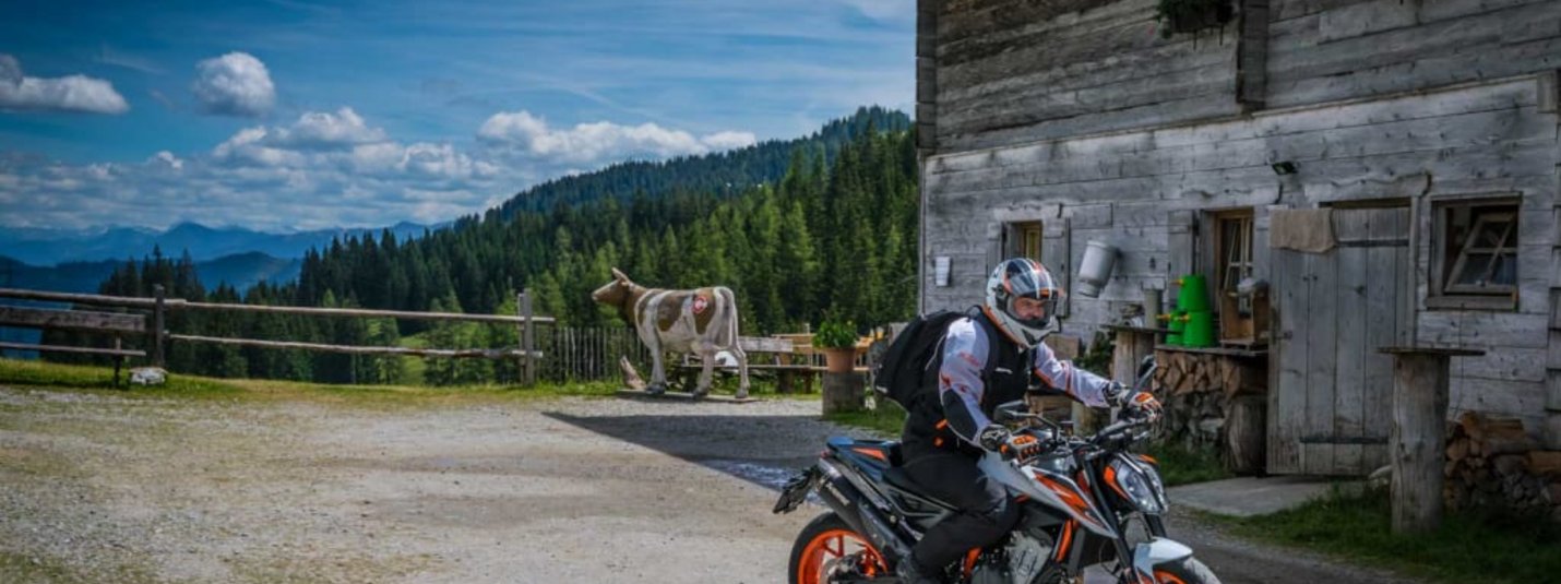 Motorradfahrer vor einer Holzhütte in ländlicher Berglandschaft mit Kuh im Hintergrund