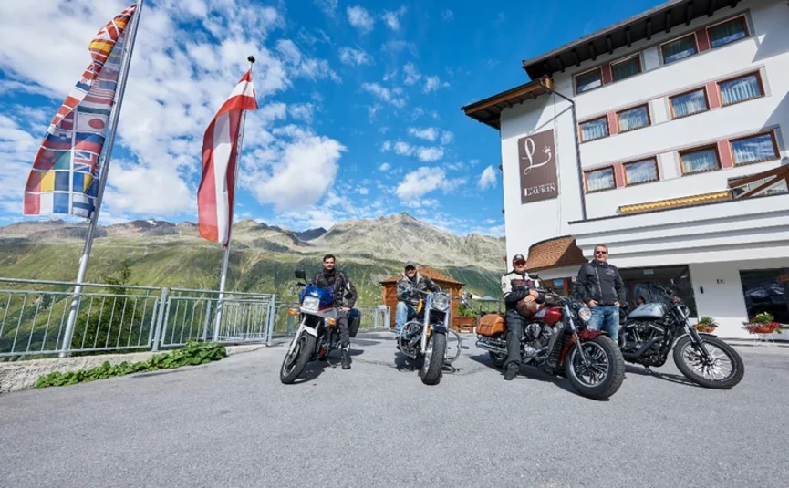 Zum Südtiroler Wein Vier Motorradfahrer vor einem Hotel mit Bergblick und Fahnen