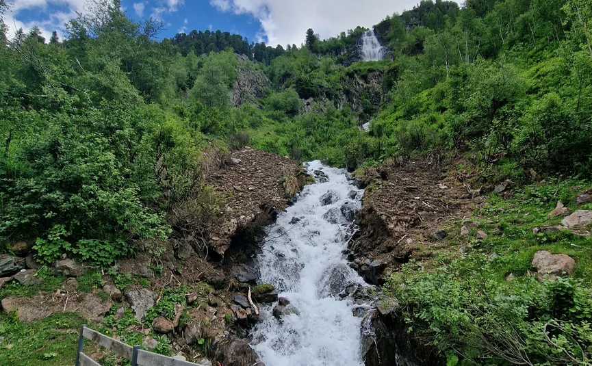 Mountain waterfall with wooden railing and forested hills under cloudy sky