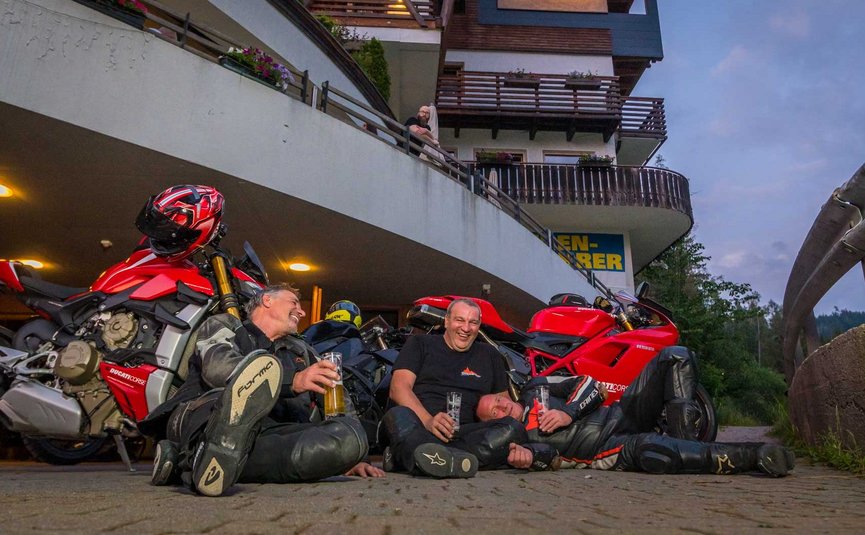 Drei Motorradfahrer sitzen lachend vor roten Motorrädern mit Getränken