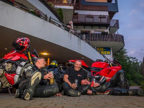 Three motorcyclists sitting on ground laughing with drinks near red motorcycles