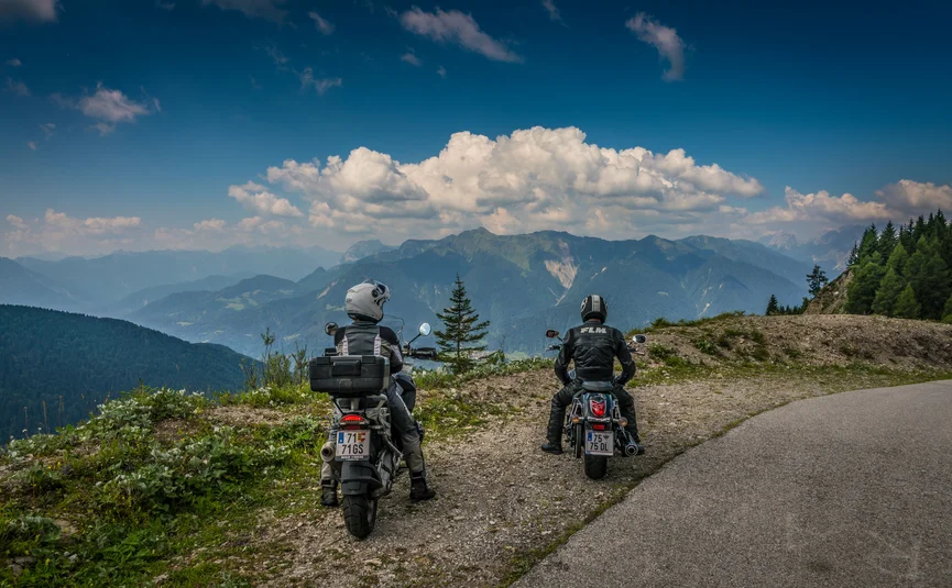 Two motorcyclists stopped on a mountain road overlooking the Alps