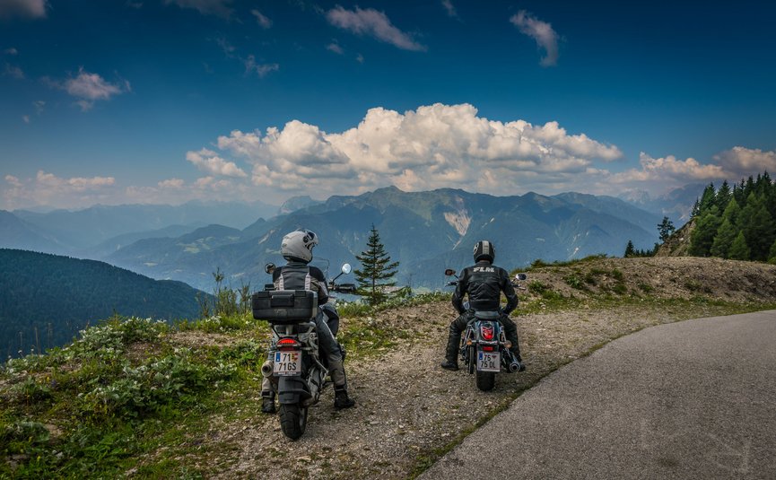 Zwei Motorradfahrer auf einem Bergweg mit Blick auf die Alpen