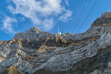 Mountain with rocky cliffs, green tree, cable car lines under blue sky