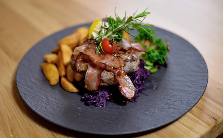 Grilled meat with bacon, potato wedges, and herbs on a black plate