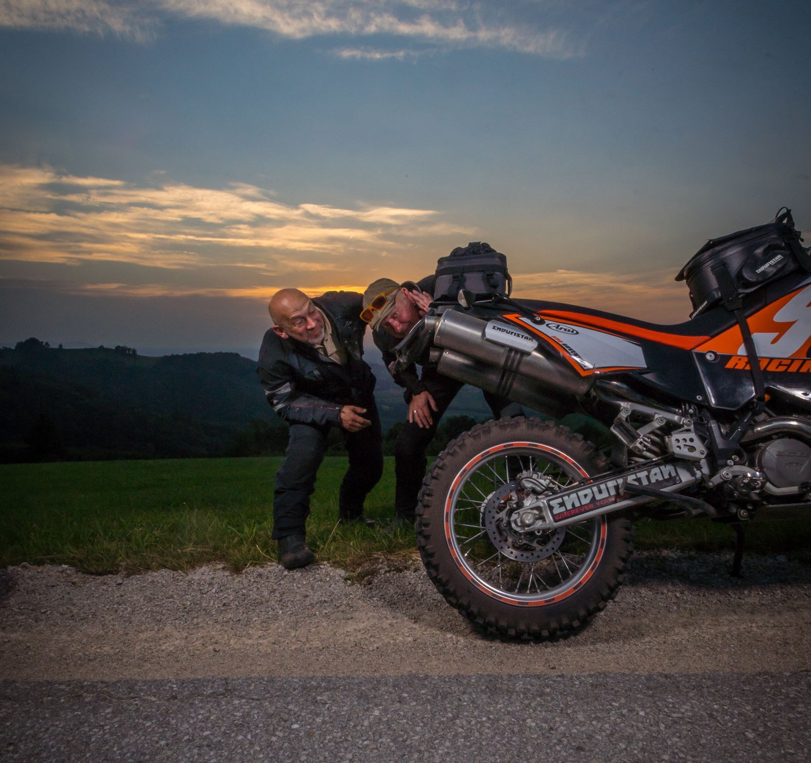 Zwei Männer schauen hinter ein Motorrad am Straßenrand bei Sonnenuntergang