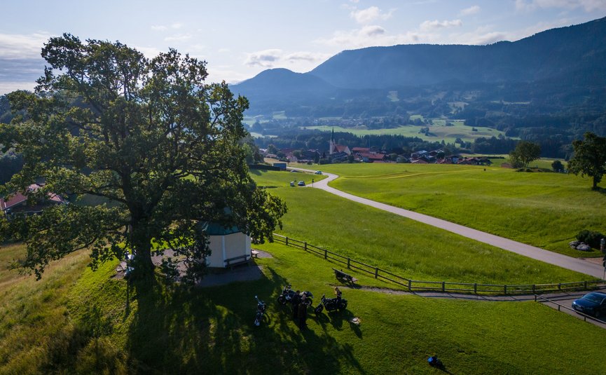 Motorradurlaub in der Region Rosenheim - Oberbayern © Moppetfoto.de Fahrradfahrer am großen Baum auf grünem Hügel mit Dorf und Bergen im Hintergrund