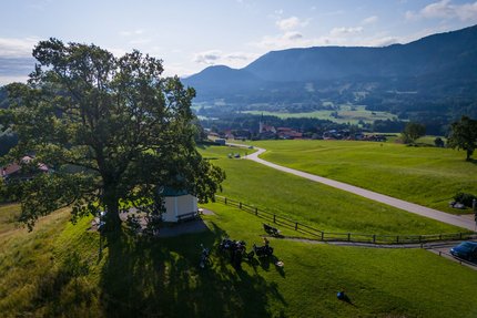 Motorrijders bij grote boom op groene heuvel met dorp en bergen op achtergrond