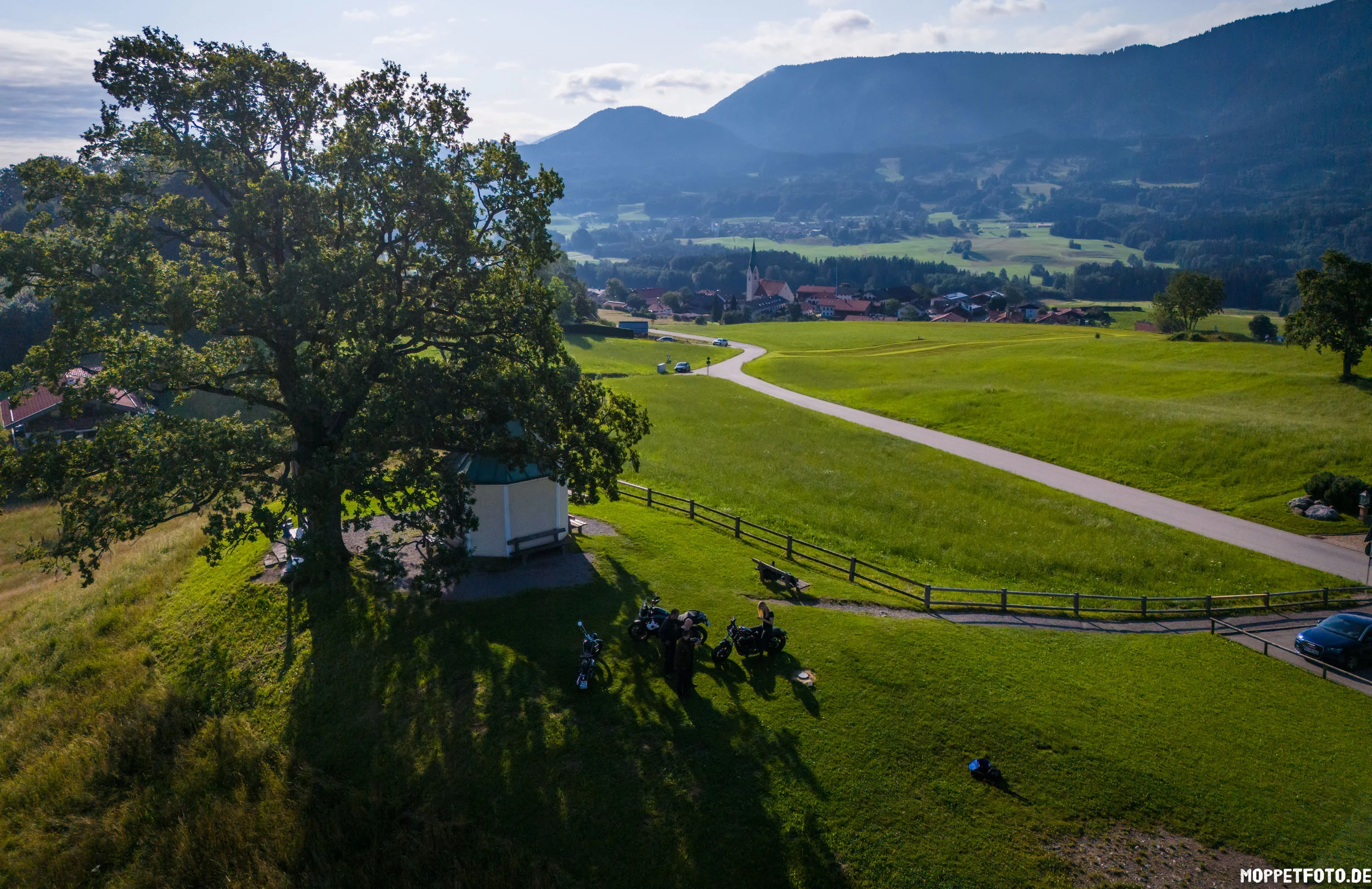 Fahrradfahrer am großen Baum auf grünem Hügel mit Dorf und Bergen im Hintergrund