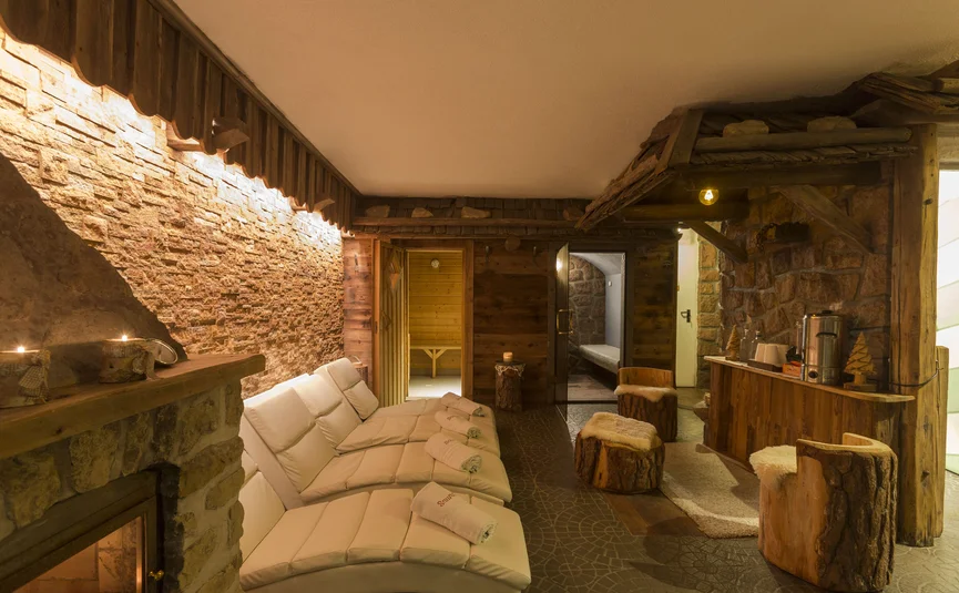 Hotel La Stüa **** © Hotel La Stüa Gemütlicher Wellnessraum mit Kamin, weißen Liegen und rustikalen Holzmöbeln