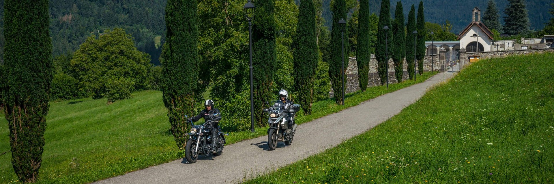 Friaul – Karnien © Moppetfoto.de Zwei Motorradfahrer fahren auf einem Weg mit Bäumen und Bergen im Hintergrund
