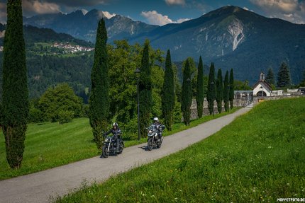 Zwei Motorradfahrer fahren auf einem Weg mit Bäumen und Bergen im Hintergrund