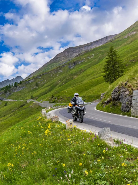 Motorcyclist riding on winding mountain road with green hills and snowy peaks