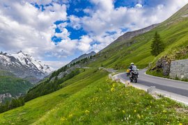 Motorradfahrer auf kurvenreicher Bergstraße mit grünen Wiesen und schneebedeckten Gipfeln
