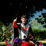 Mann mit Motorradkleidung zeigt Daumen hoch neben rotem Motorrad im Freien