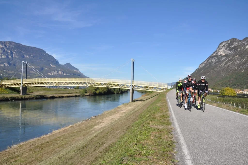 Group of cyclists on path next to river and bridge in mountainous area