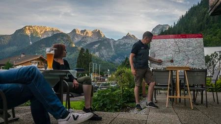 Man wijst op wandelkaart met bergen op de achtergrond, twee personen zitten met drankjes