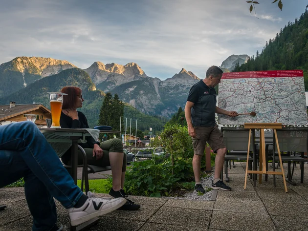 Man wijst op wandelkaart met bergen op de achtergrond, twee personen zitten met drankjes