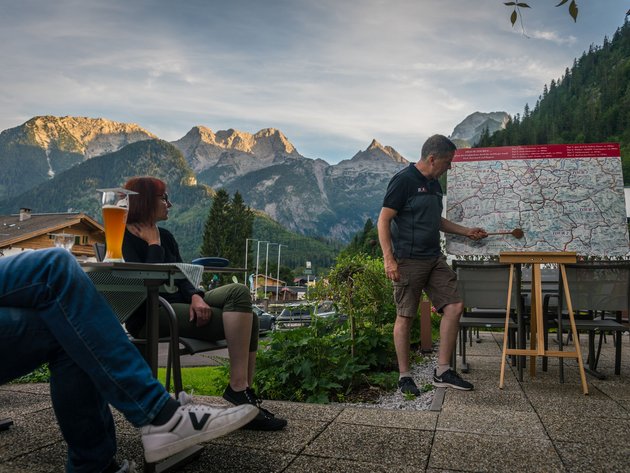 Qualitätsversprechen © Moppetfoto.de Mann zeigt auf Wanderkarte vor Bergkulisse, zwei Personen sitzen mit Getränken