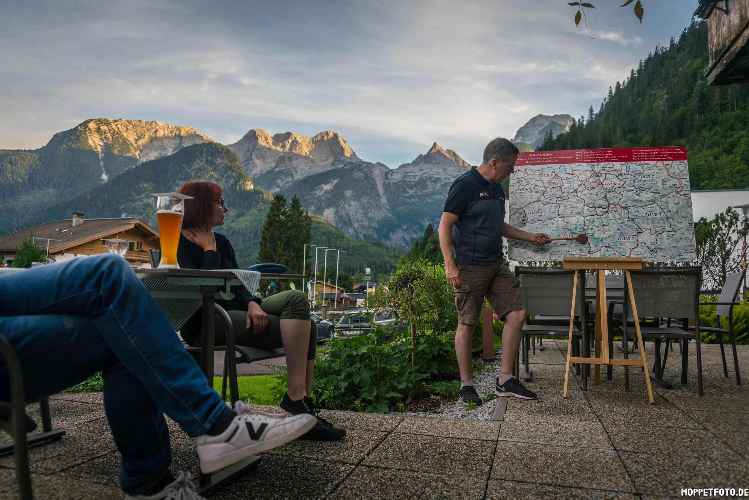 Man pointing at hiking map with mountains in background, two people seated with drinks