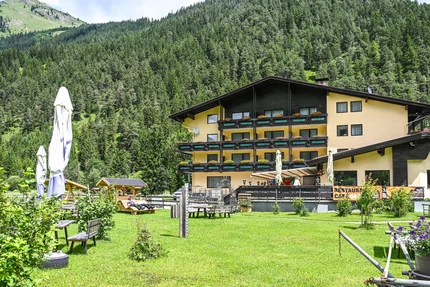 Gelbes Alpenhotel mit Garten vor bewaldetem Berg im Sommer