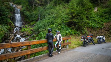 Zwei Motorradfahrer stehen auf einer Brücke mit Wasserfall und Wald im Hintergrund