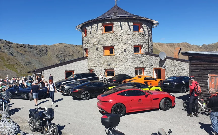 Sports cars and motorcycles near round stone building in mountains