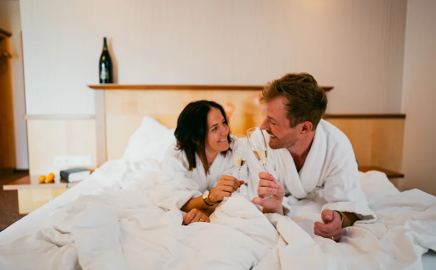 Couple in bathrobes clinking champagne glasses on bed